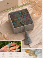 May include: A square gray box with a patterned fabric lid featuring blue, pink, and gold cloud motifs, finished with a gray tassel. The set includes a multi-colored crystal bead bracelet, a thank you card, and a box labeled Mira Luna Stone.